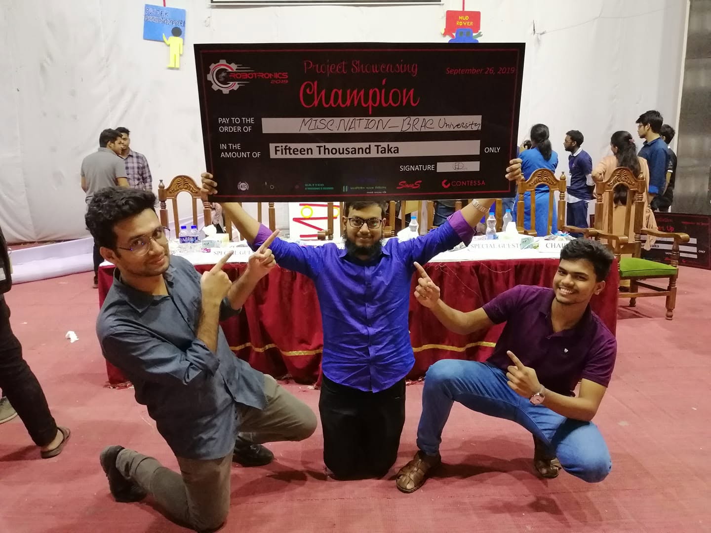 Three team members kneeling behind a Robotronics 2019 Project Showcasing Champion cheque for fifteen thousand taka.